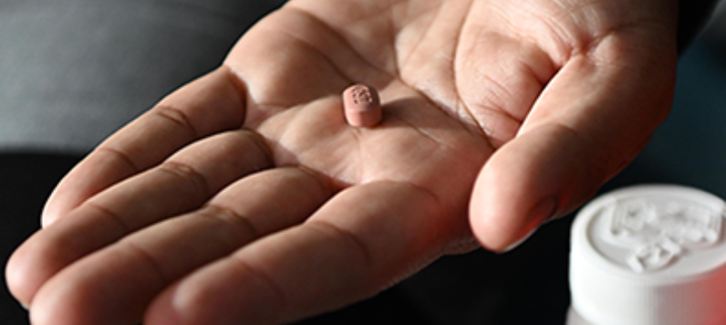 A hand holding a pill next to a pill bottle.