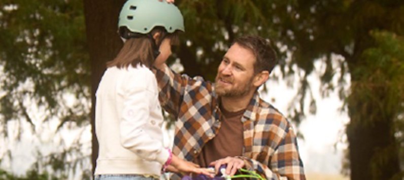 A man crouched beside his daughter on her bike.